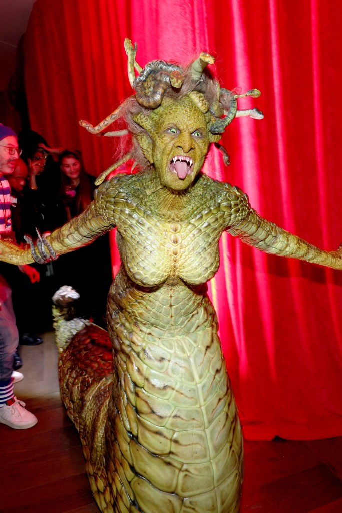 Heidi Klum in an elaborate, scaly green Medusa costume with a snake-like lower body, posing in front of a red curtain.