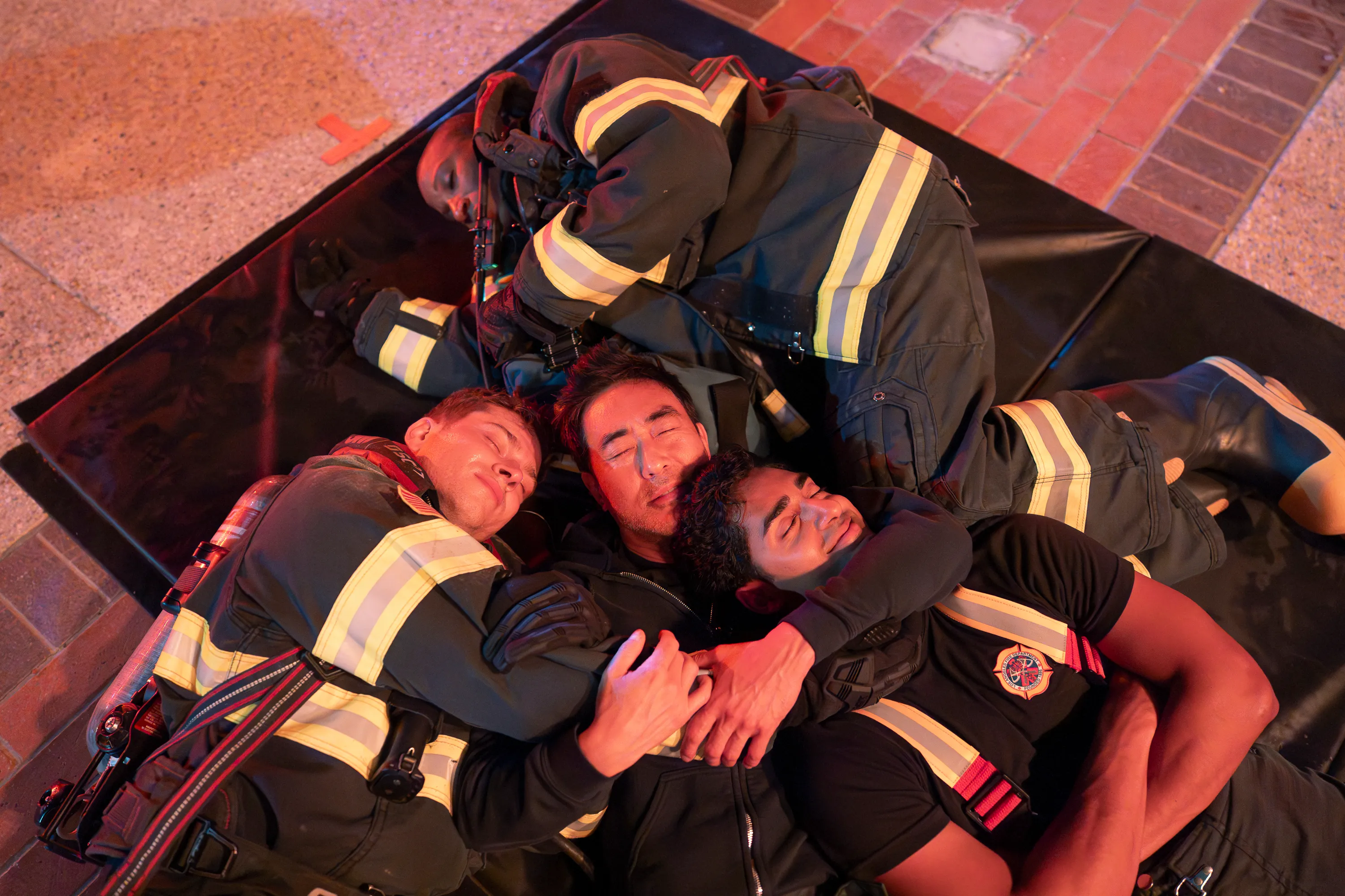 Four firefighters lie tangled together on a black rescue mat, their eyes closed as if exhausted.