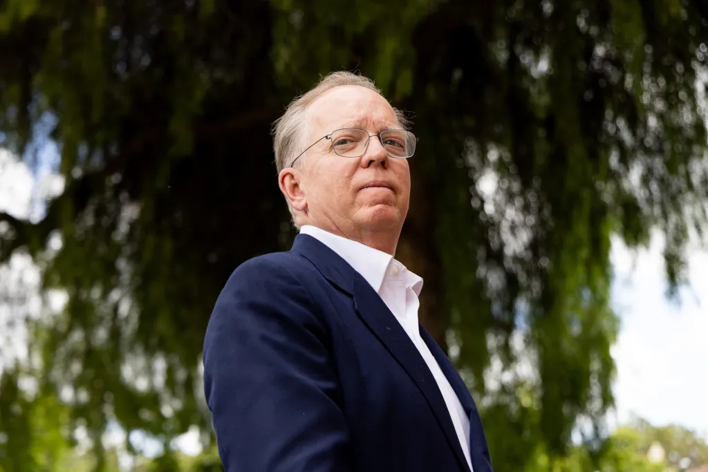 Herb Morgan, California state controller candidate, stands outdoors in a blue suit and glasses against a backdrop of trees.