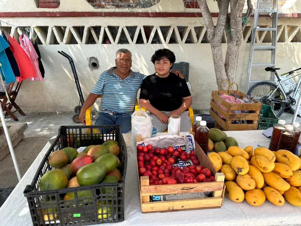Hernan Martinez, 16, sitting with an older man behind a fruit stand.