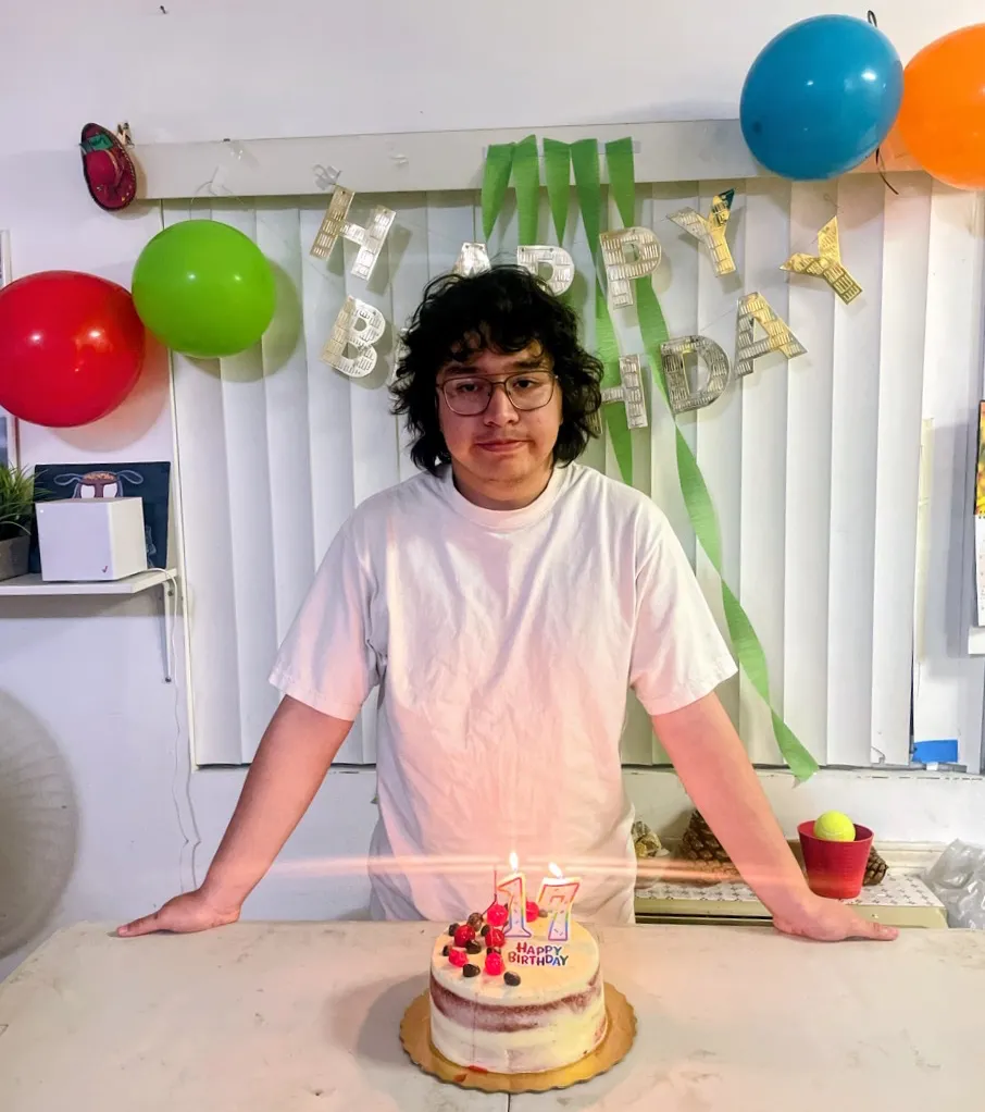 Xavier Nungarary, 17, posing behind a birthday cake with lit candles and birthday decorations in the background.