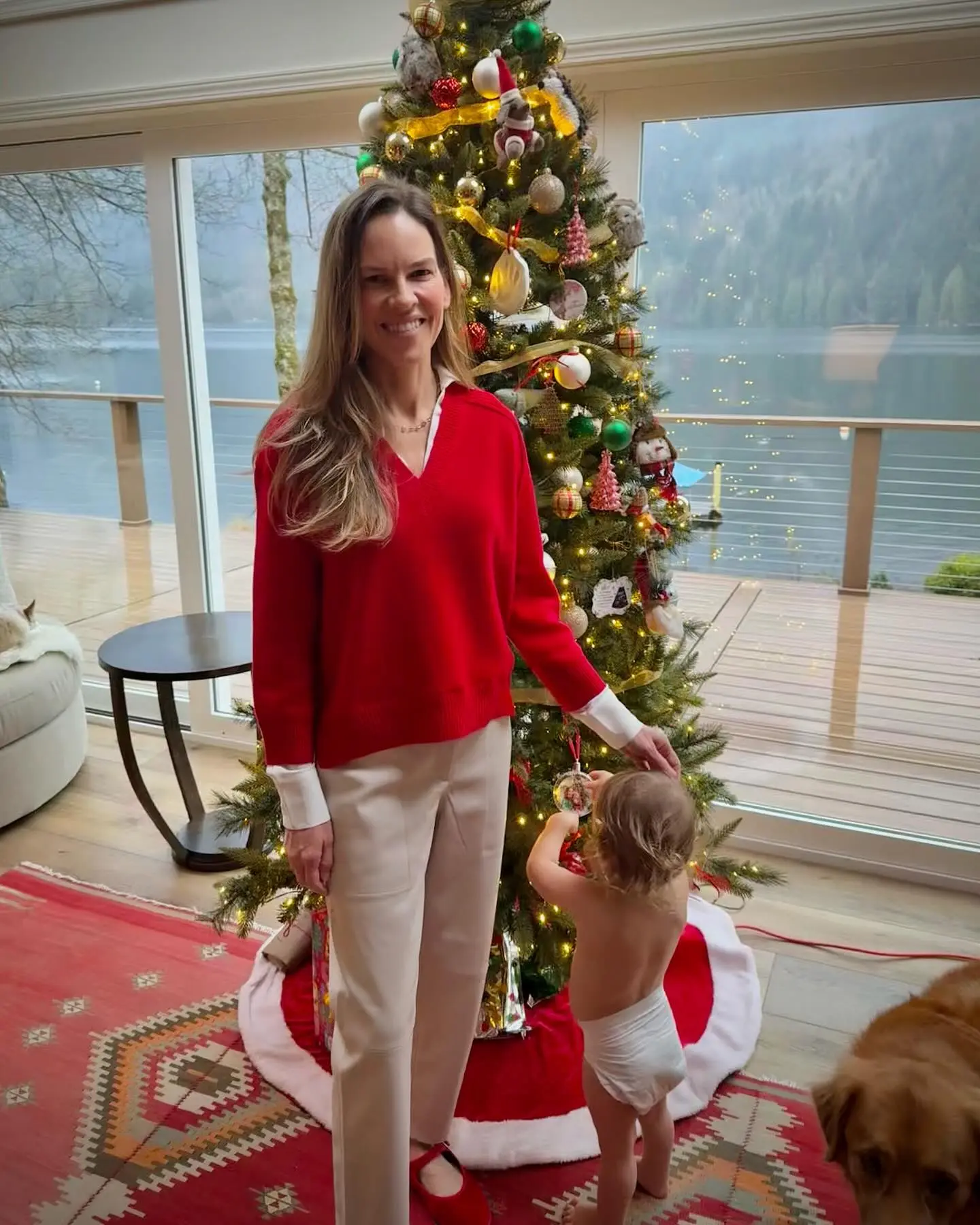 Hilary Swank stands beside a decorated Christmas tree with a toddler and a dog in front of a large window overlooking a lake.