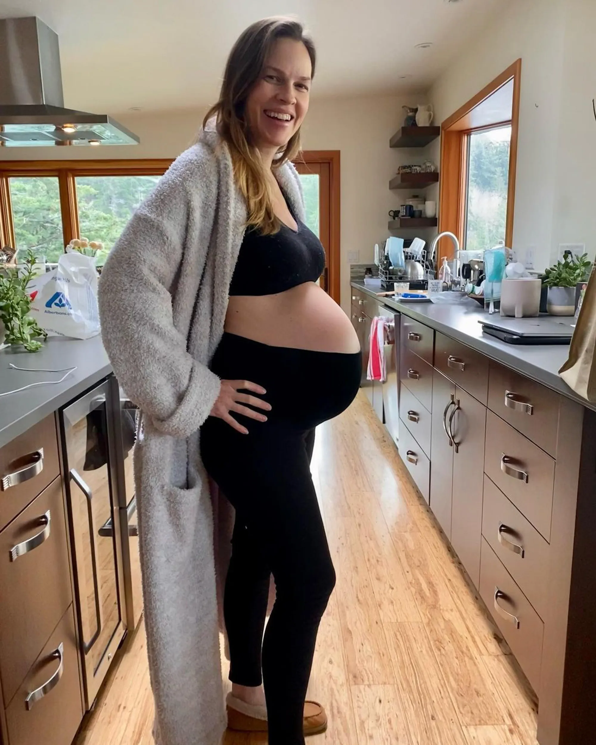 Hilary Swank stands pregnant in a kitchen, wearing a black sports bra, black leggings, and a grey robe, smiling at the camera.