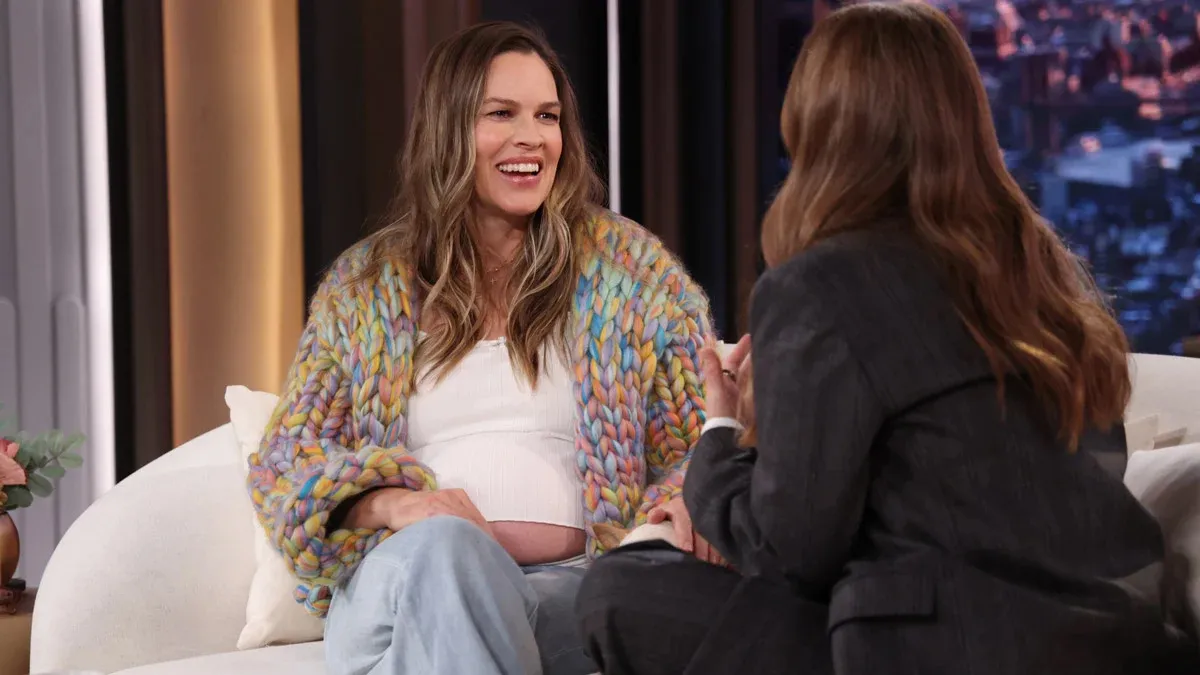 Hilary Swank, wearing a colorful chunky knit cardigan and a white top, sits on a couch while talking to Drew Barrymore on her talk show.