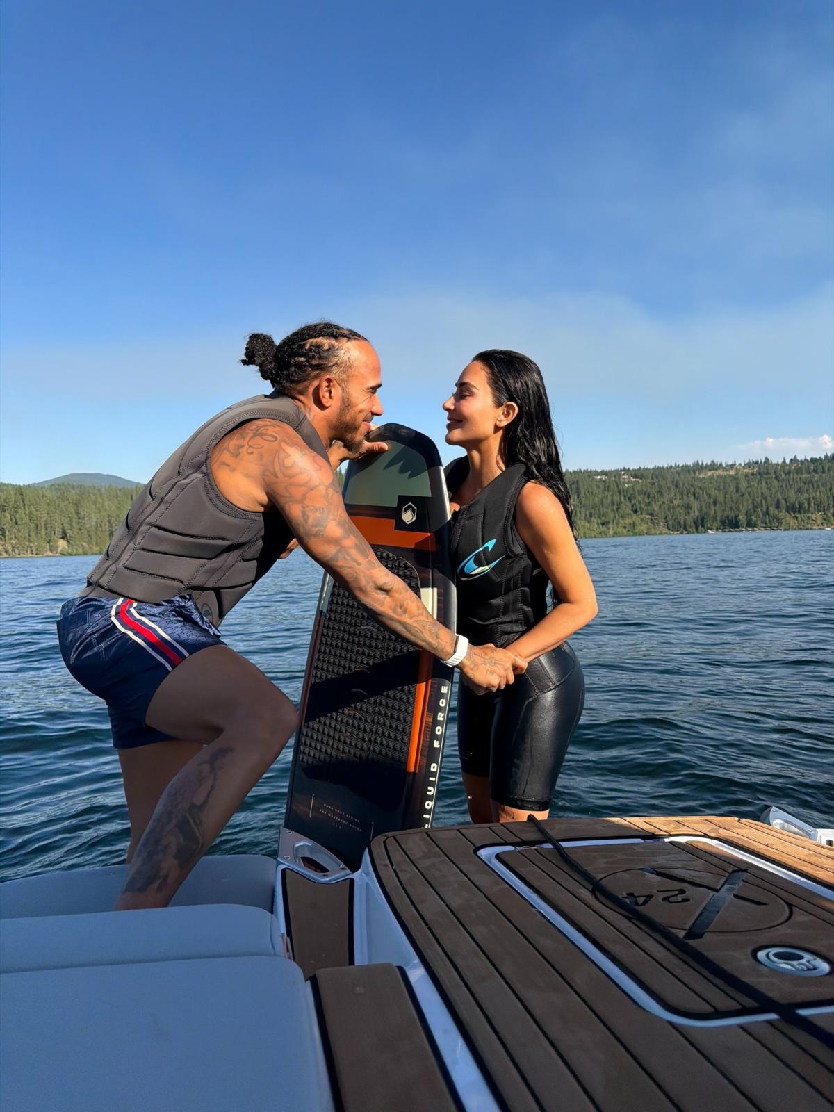 Lewis Hamilton and Kim Kardashian standing together on a boat holding a wakesurf board, smiling at each other.