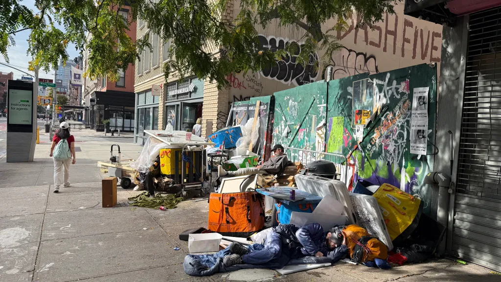 A homeless encampment with piles of belongings and a person lying on the sidewalk on a city street.