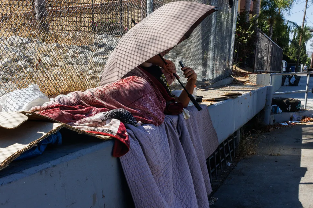 A homeless man lies under an umbrella and blankets on a concrete ledge in a cul-de-sac.