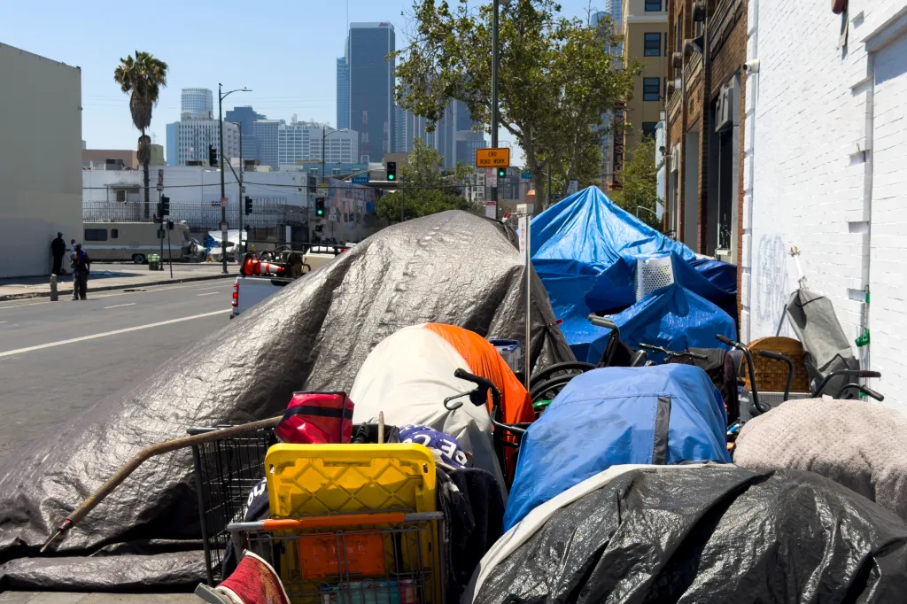 Tents and makeshift shelters line the sidewalk on Skid Row in Los Angeles, with the city's skyline visible in the background.