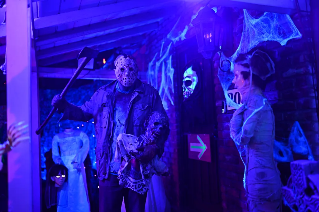 Homeowner Diego Linares, dressed as Jason from Friday the 13th, holding an axe and a prop head, walks through his Halloween-decorated house.