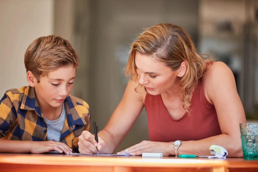 A mother helps her young son with his homework, guiding his hand as he writes on a paper at a table.