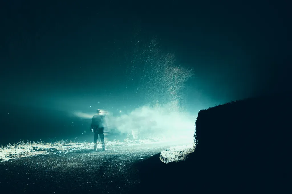 A man vanishes into smoke in front of bright lights on a country road on a foggy winter night in a horror, sci-fi concept photo.