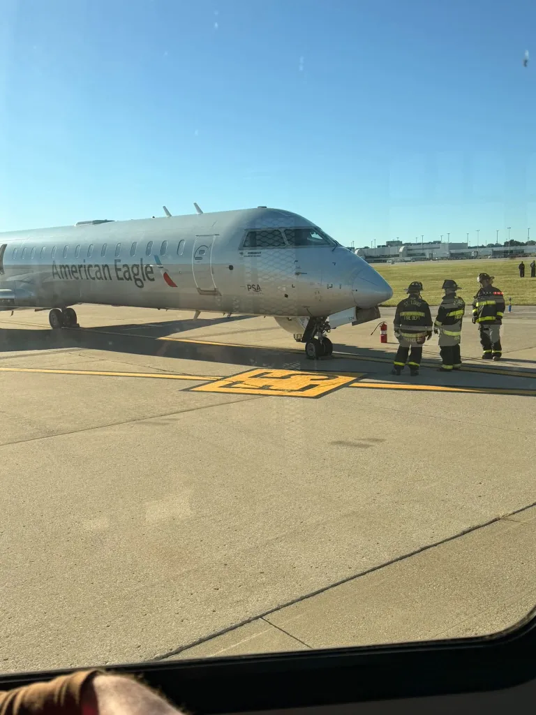 An American Eagle plane parked on an airfield with three firefighters standing near the nose.