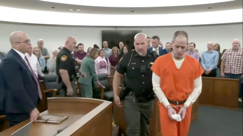 Caleb Flynn, in an orange jumpsuit, exits the courtroom accompanied by a law enforcement officer.