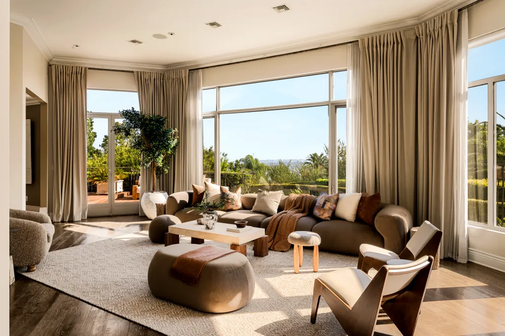 An interior view of Dolly Parton's home in Los Angeles, featuring a spacious living room with large windows overlooking outdoor greenery and a seating area with a sofa, armchairs, and a coffee table.