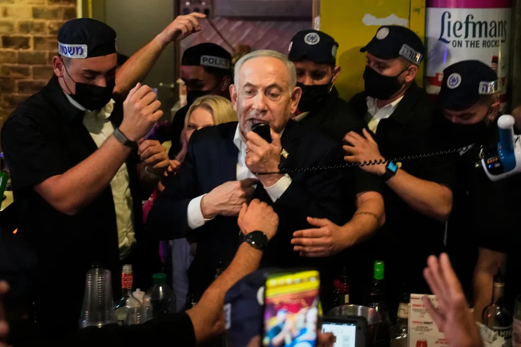 Israeli Prime Minister Benjamin Netanyahu speaks into a microphone, surrounded by security personnel and supporters.