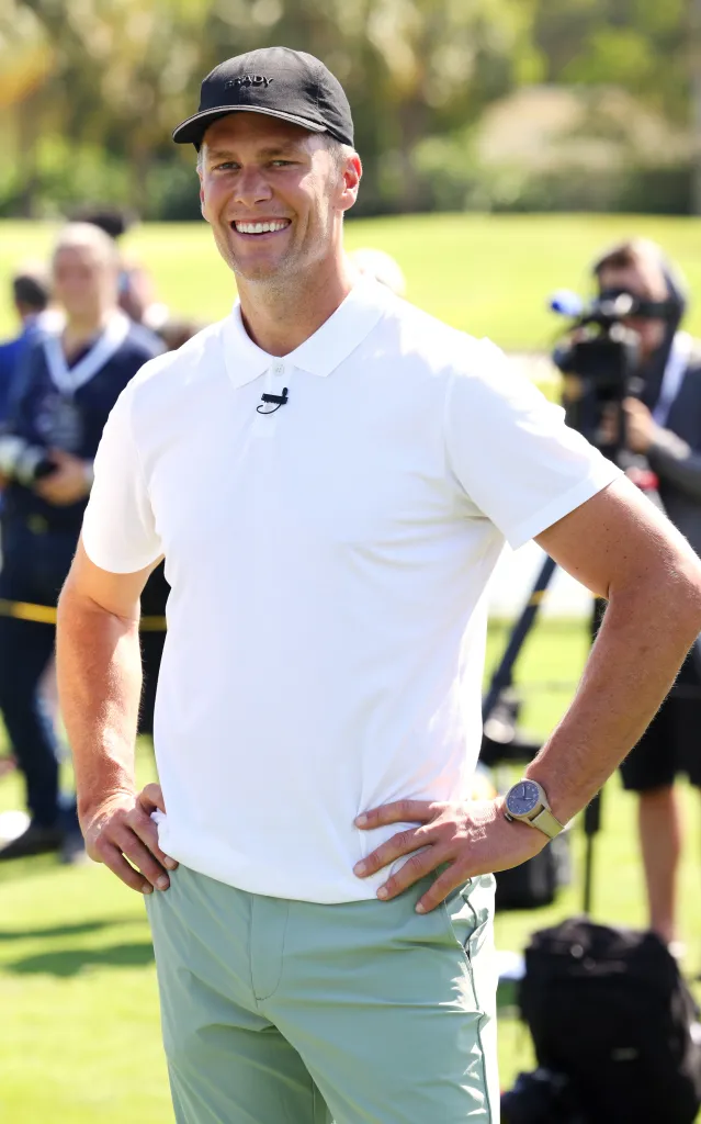 Tom Brady wearing a white polo shirt, black cap, and a watch at a golf event.