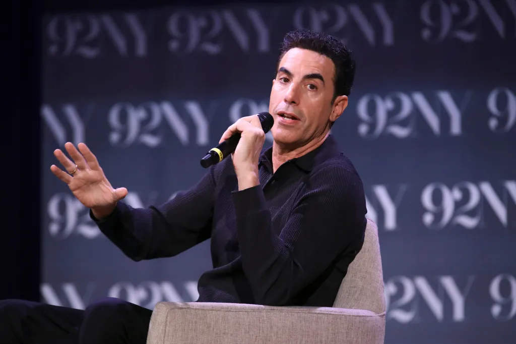 Sacha Baron Cohen speaks into a microphone while seated on stage at The 92nd Street Y.