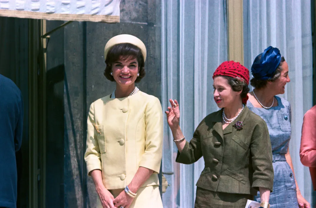 Jacqueline Kennedy, wearing a yellow dress and matching hat, stands with dignitaries during a state visit to France.