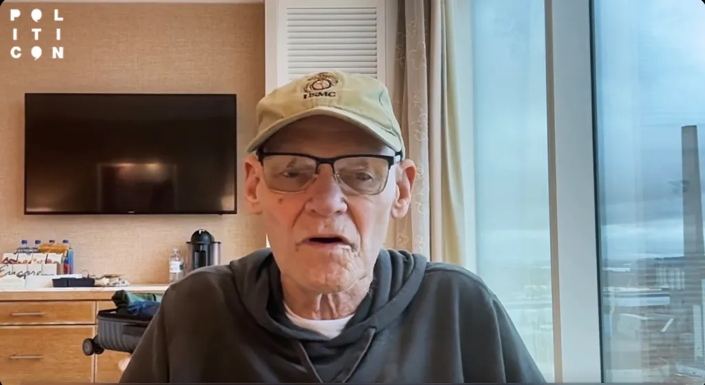James Carville wearing a cap and glasses while speaking indoors.
