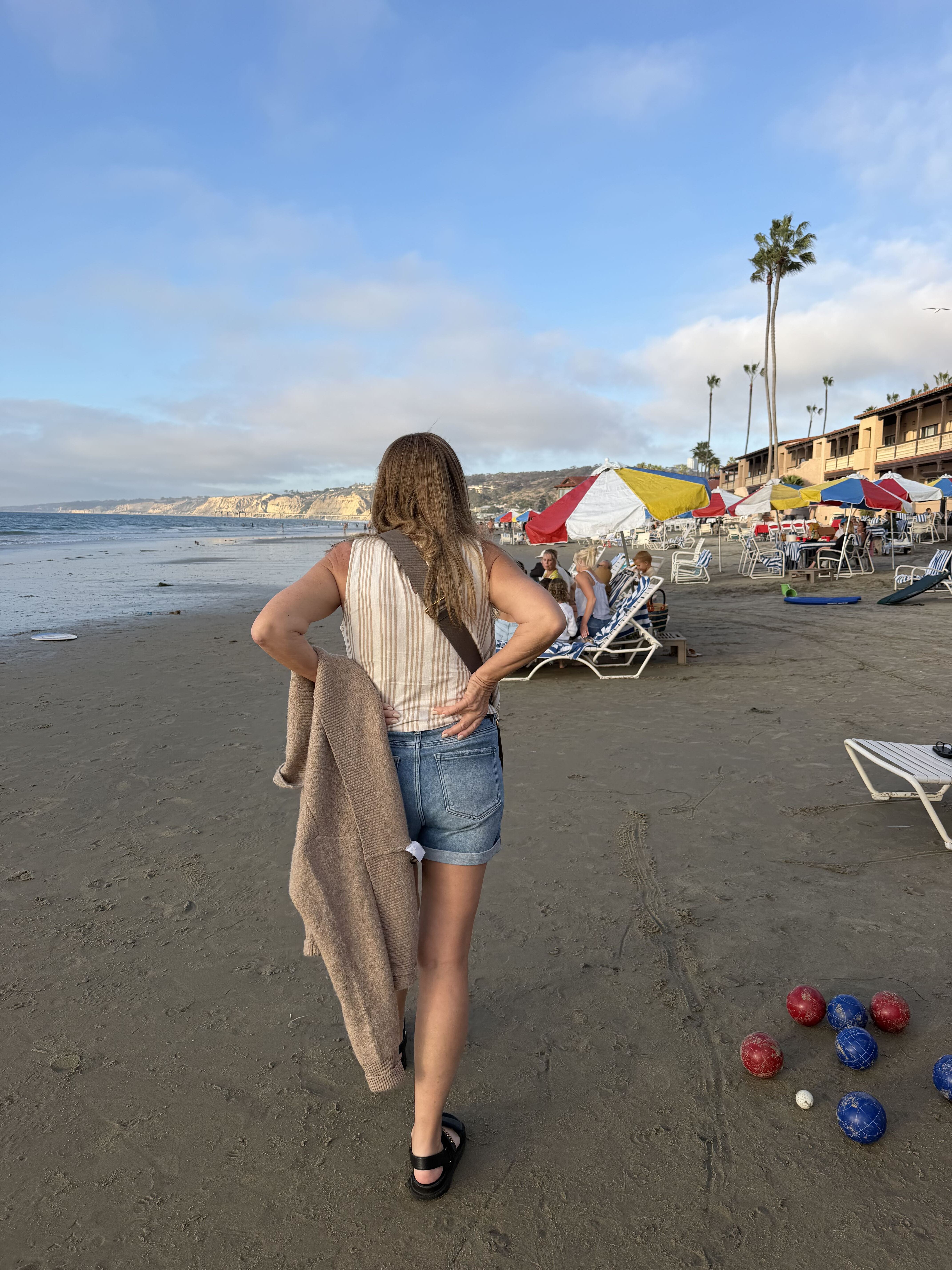 Jeana Keough stands on a beach, facing away from the camera, holding a sweater.
