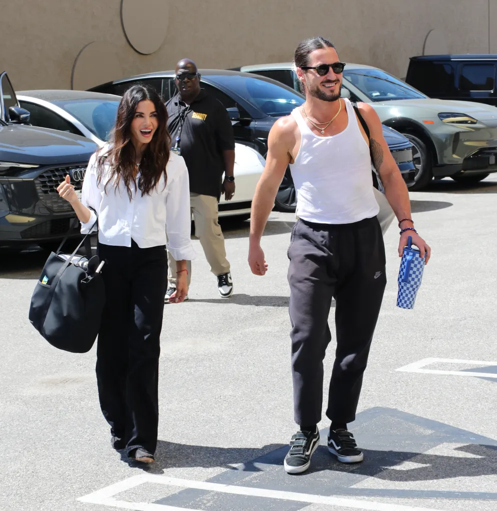 Jenna Dewan and Val Chmerkovskiy walk together outdoors in a parking lot, with Dewan in a white shirt and black pants holding a bag, and Chmerkovskiy in a white tank top and dark pants holding a water bottle.