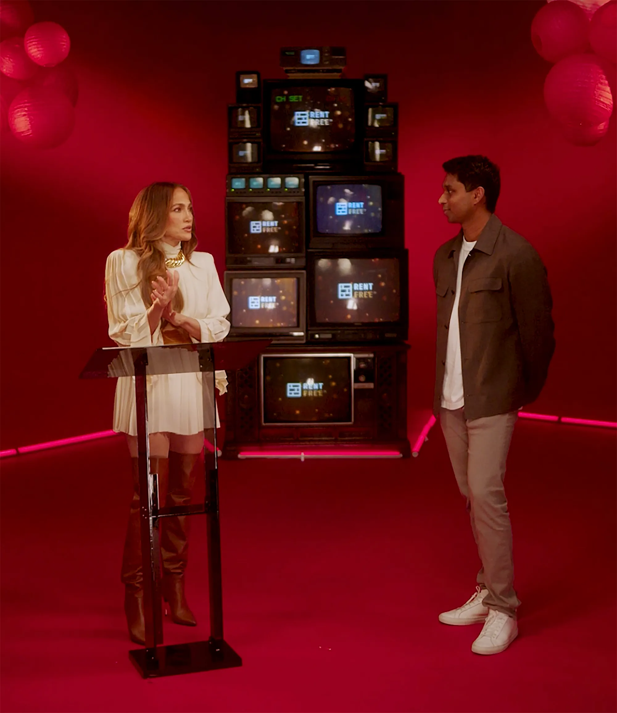 Jennifer Lopez and Ankur Jain standing in a red room with a stack of televisions behind them.