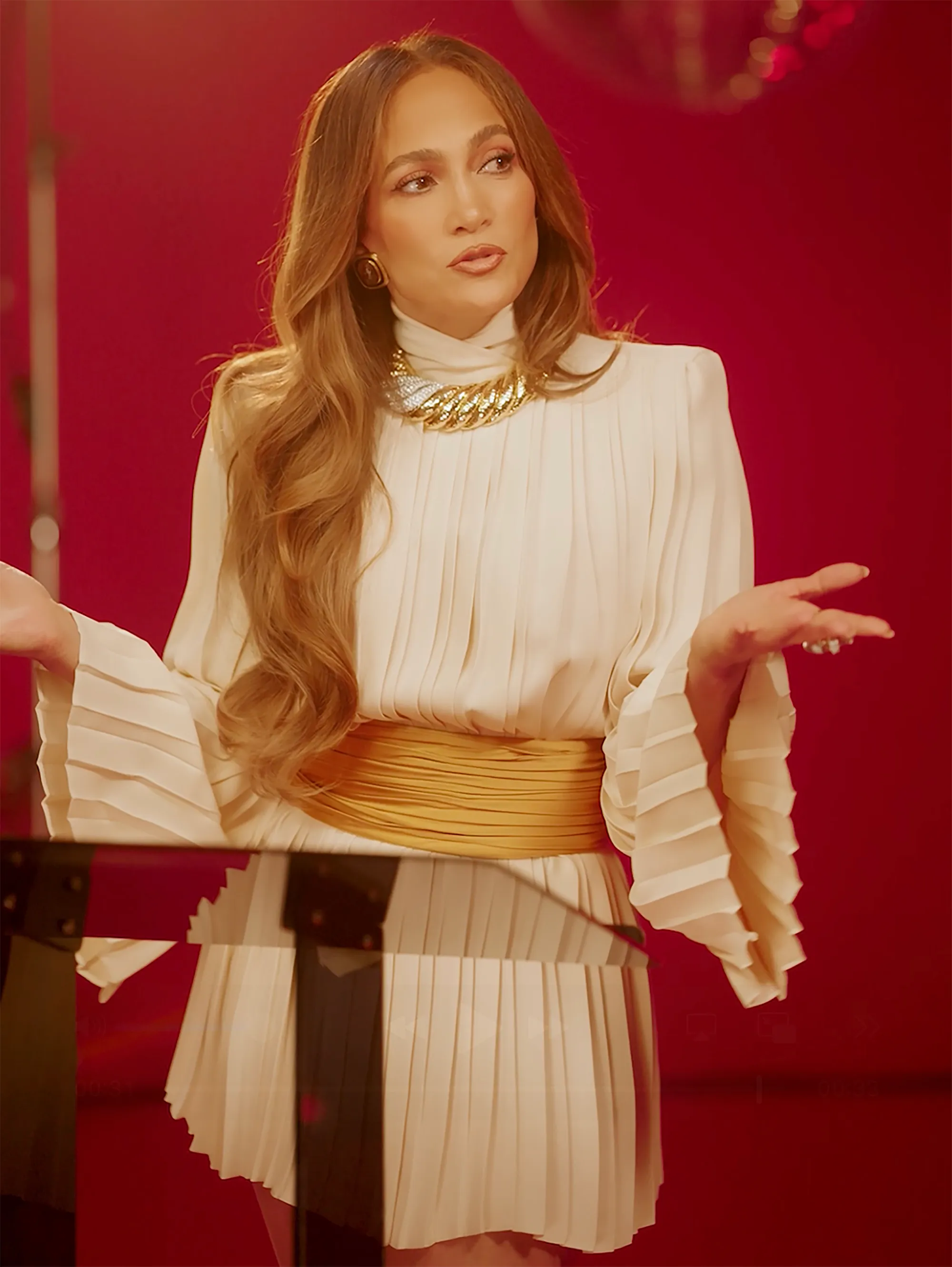 Jennifer Lopez wearing a cream-colored, pleated dress with a gold belt and necklace, standing behind a podium against a red background.