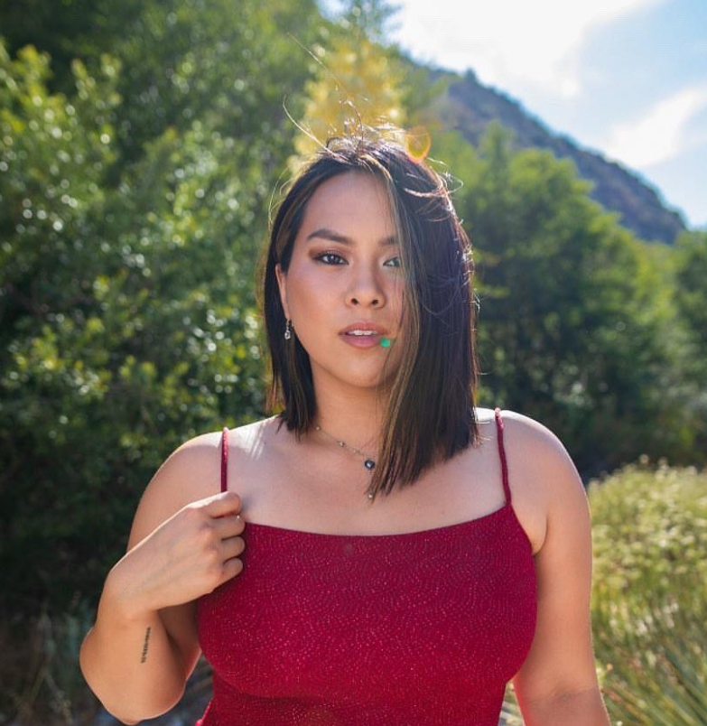 Fitness coach and personal trainer Jenny Voo poses outdoors in a red dress with a natural, leafy background.