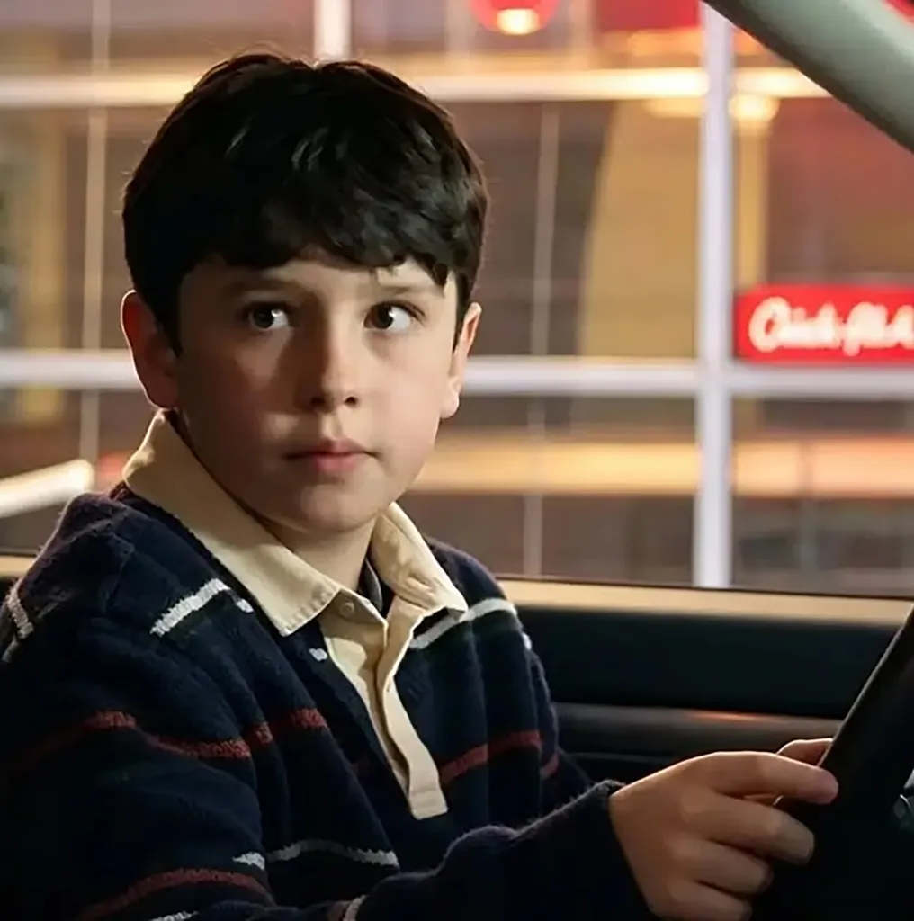 A young boy with dark hair sitting in the driver's seat of a car, looking out the window.