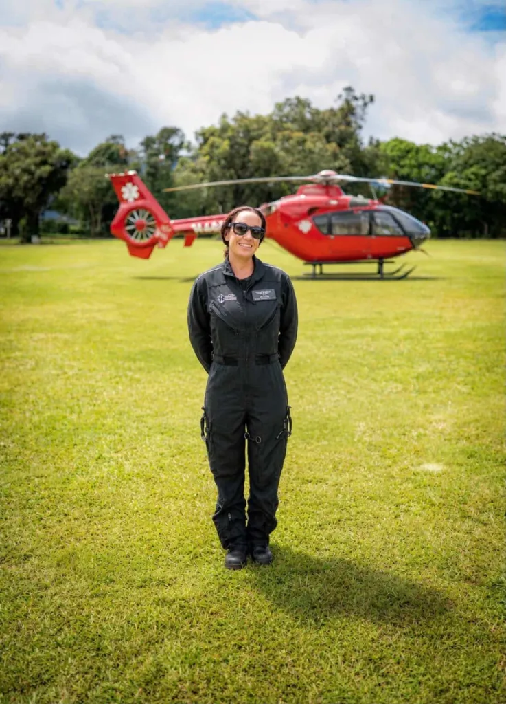 Jessica DeVries stands in a flight suit in front of a red medical helicopter on a grassy field.