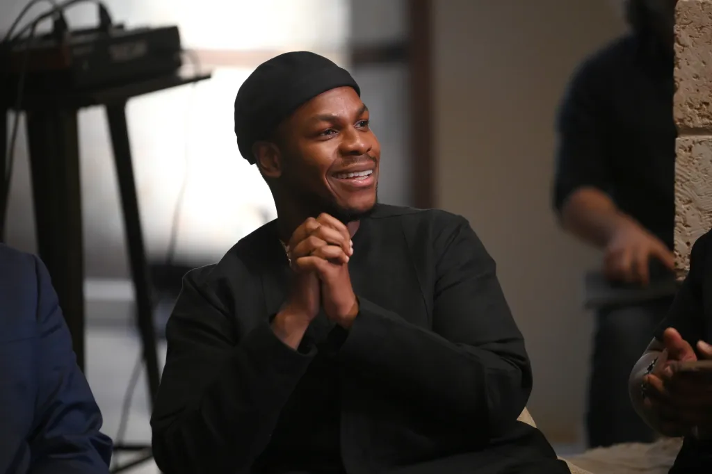 John Boyega smiling with hands clasped during a round table discussion.
