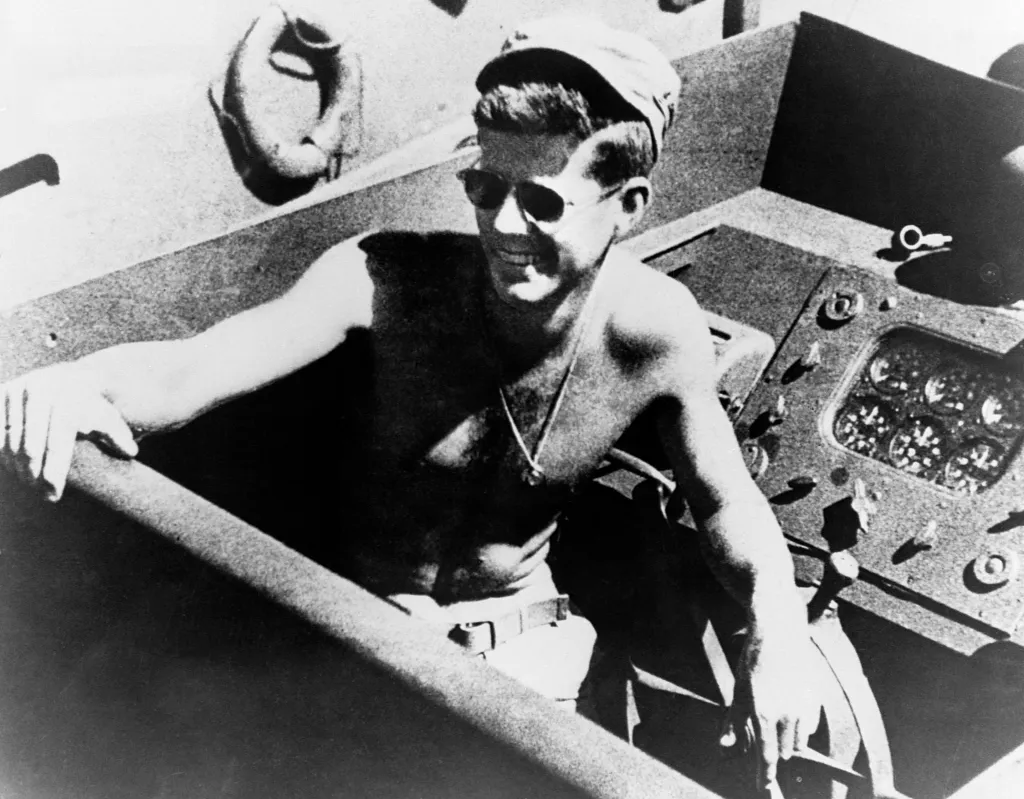 John F. Kennedy smiling shirtless in a U.S. Navy PT boat, wearing sunglasses and a cap.