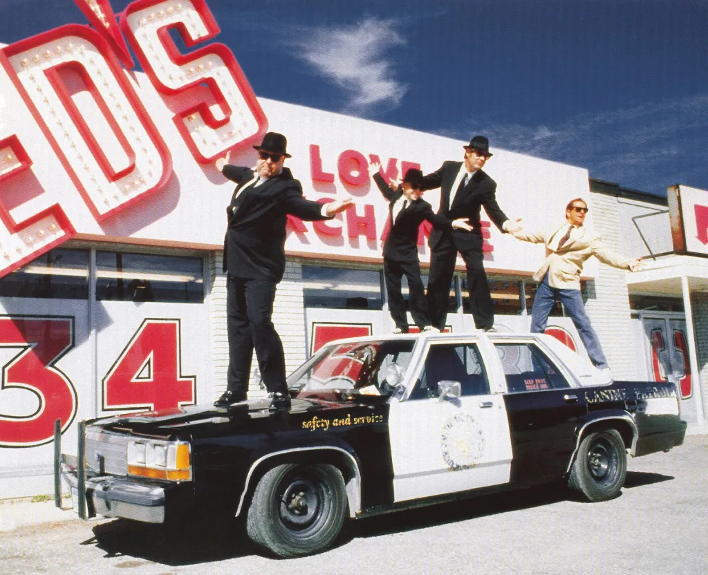 John Goodman, J. Evan Bonifant, Dan Aykroyd, and Jonny Lang pose on a police car in a scene from the movie Blues Brothers 2000.