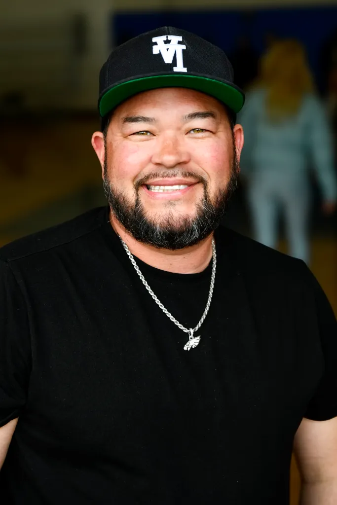 Jon Gosselin smiling and wearing a black t-shirt, a silver necklace, and a black baseball cap.