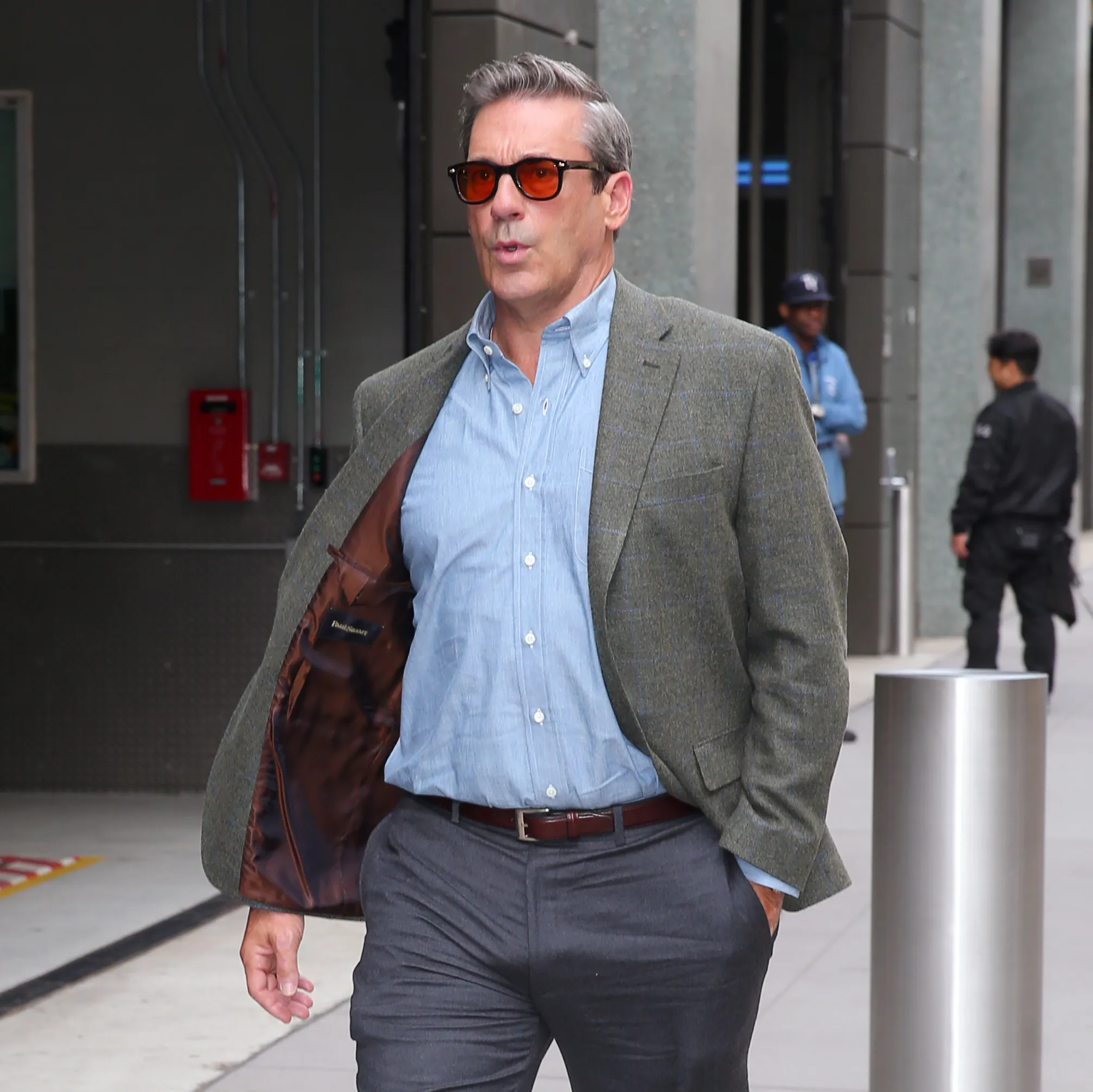 Jon Hamm walks on a street in a green blazer, blue button-down shirt, and sunglasses.