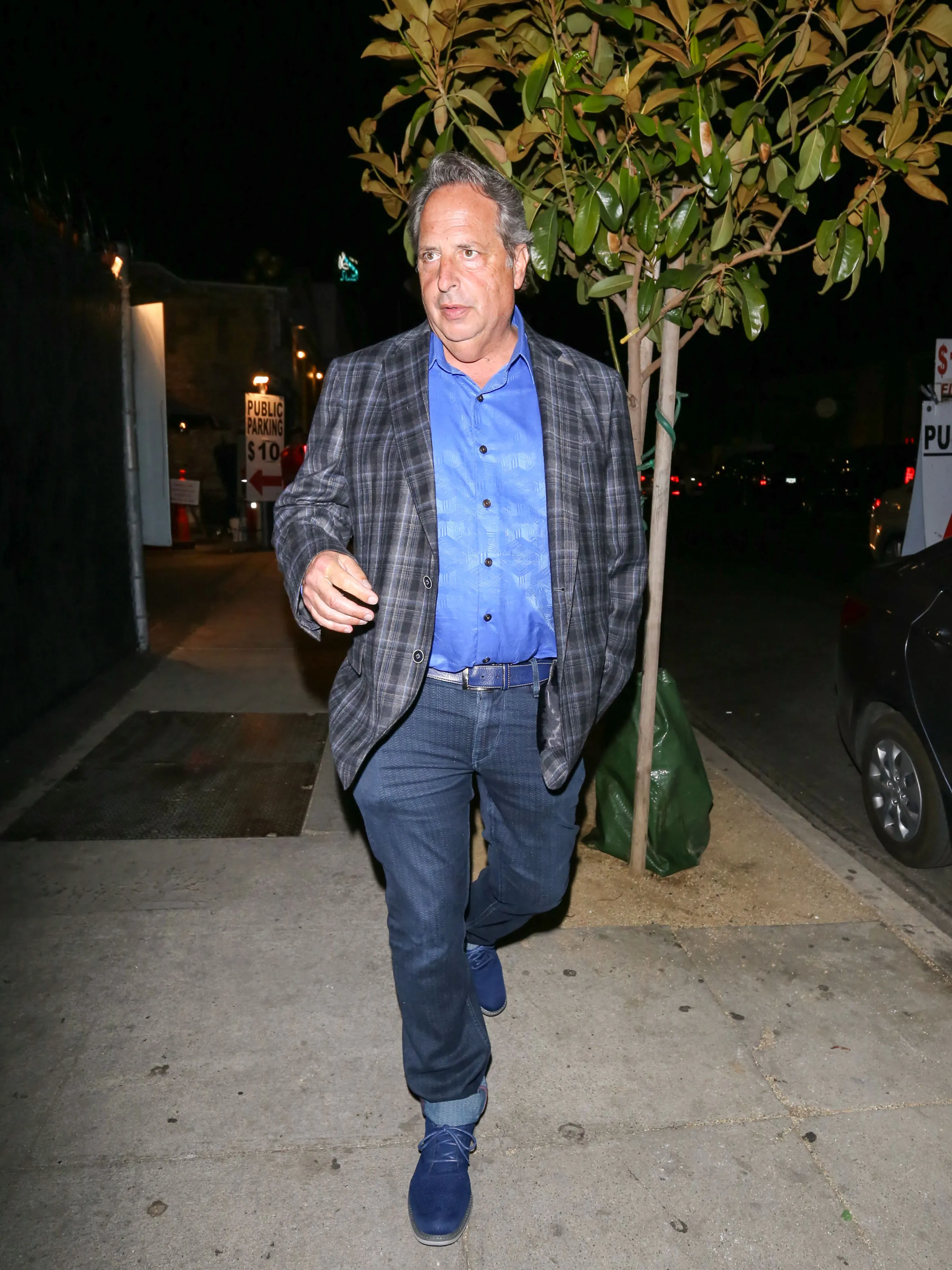 Jon Lovitz walks down a city sidewalk at night wearing a plaid blazer, blue shirt, and jeans.