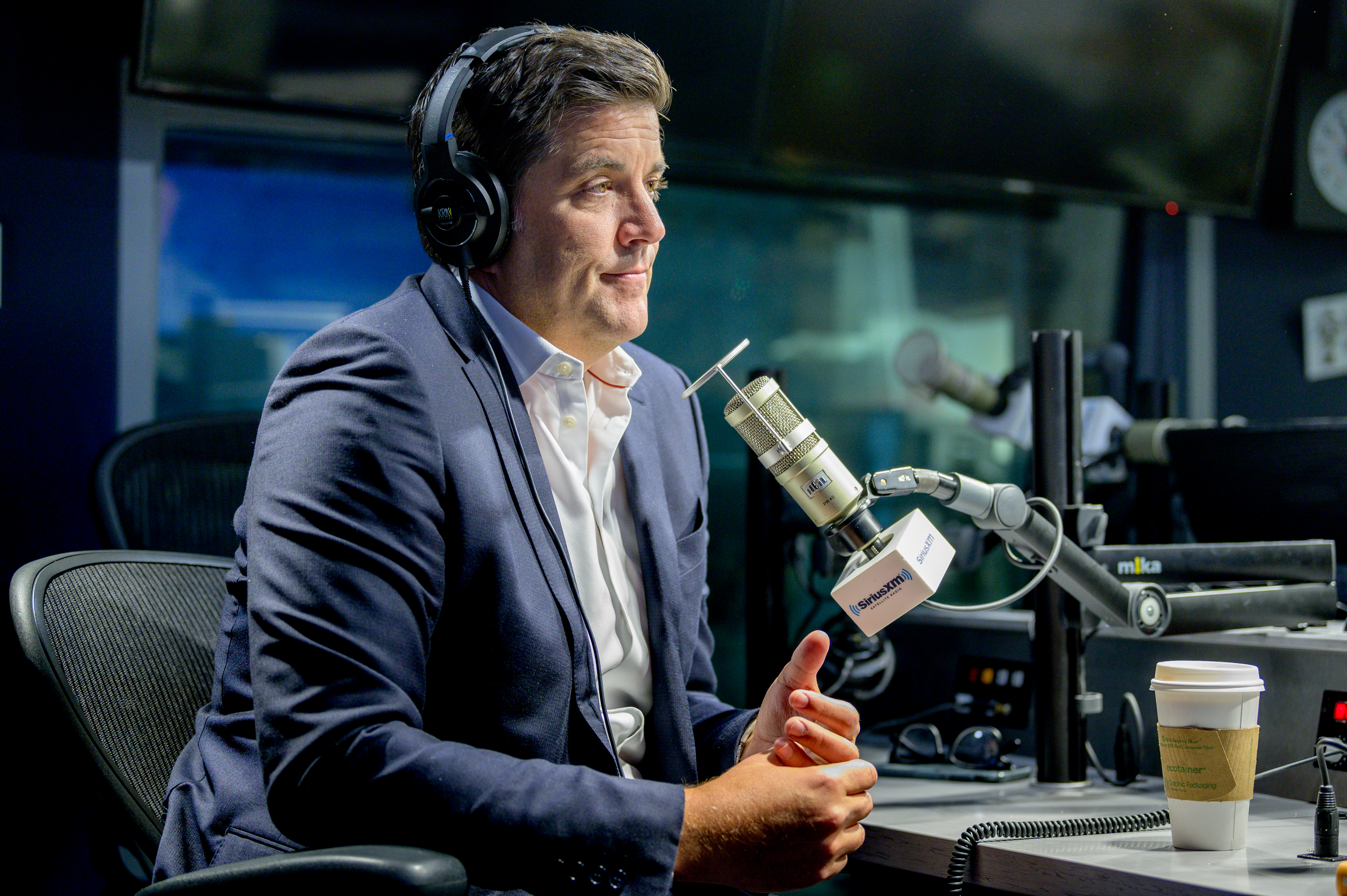 Journalist Josh Elliott wearing headphones and a suit, sitting in a studio in front of a microphone.