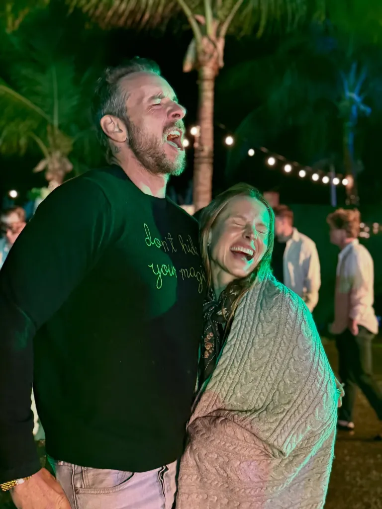 Dax Shepard and Kristen Bell laughing together outdoors at night under string lights.
