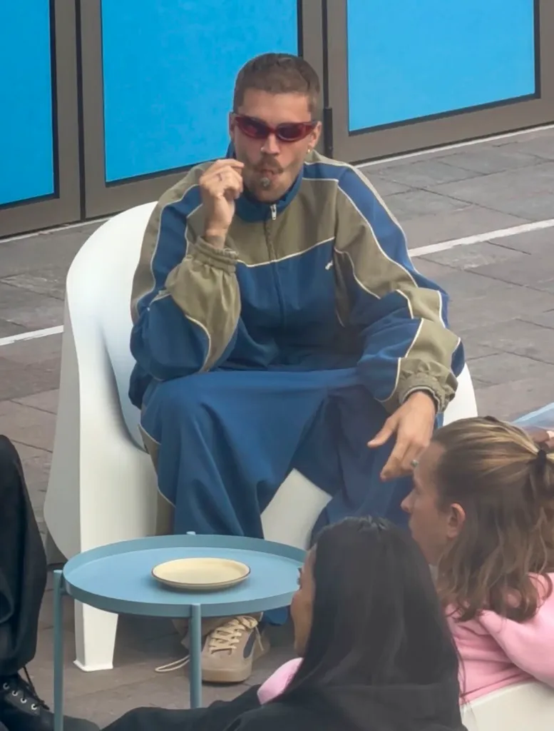 Justin Bieber sits outdoors in a blue tracksuit and sunglasses, smoking a cigar while chatting with friends.