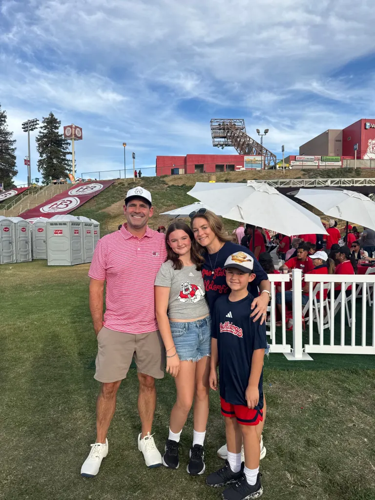 Justin Derby and his family stand together outdoors at an event, with stadium seating and white umbrellas in the background.