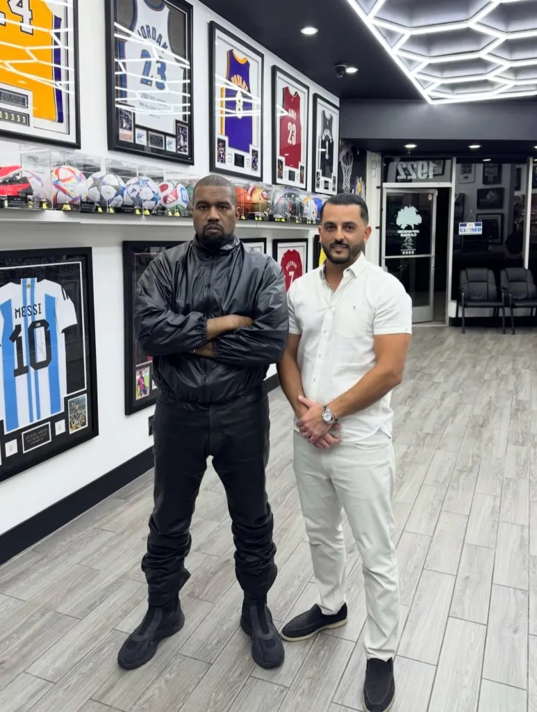 Kanye West standing with his arms crossed in a sports memorabilia store, next to another man in a white shirt.
