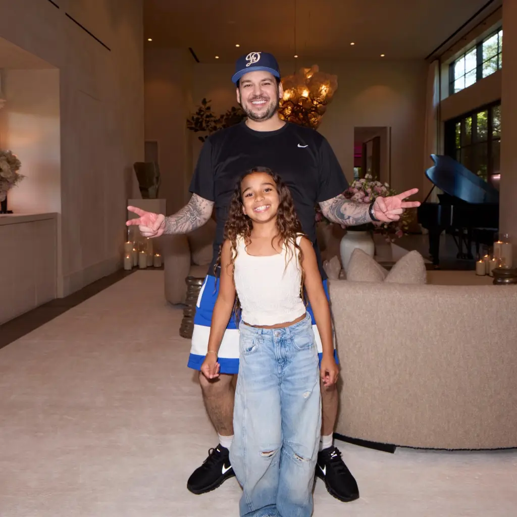 Rob Kardashian and his daughter standing together in a living room, both smiling and flashing peace signs.