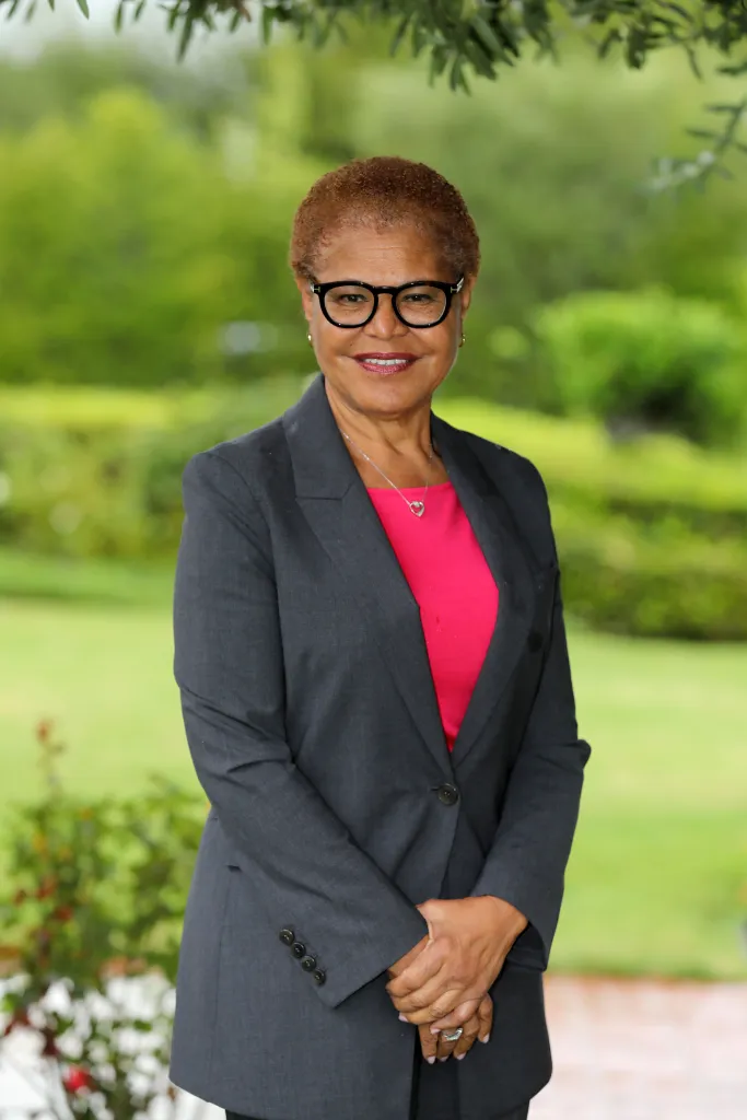 Karen Bass wearing a grey suit and pink shirt, smiling for the camera outdoors.