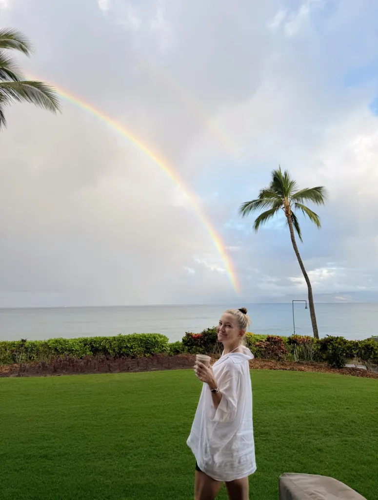 Kate Hudson holds a cup while posing in front of a double rainbow over the ocean during a tropical getaway.