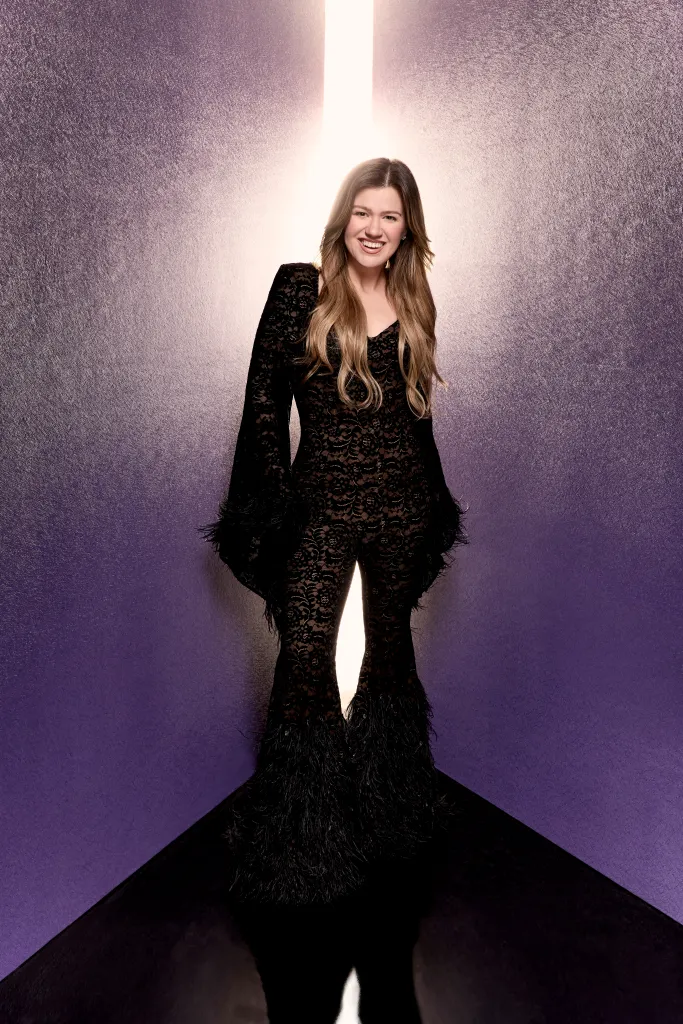 Kelly Clarkson in a black lace jumpsuit standing against a purple backdrop.