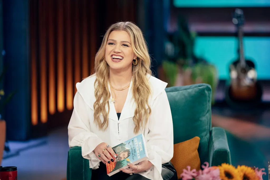 Kelly Clarkson smiles while holding a book on her talk show set.