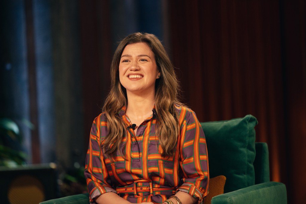 Kelly Clarkson sits in a green armchair on the set of her talk show, wearing a colorful patterned blouse and smiling.