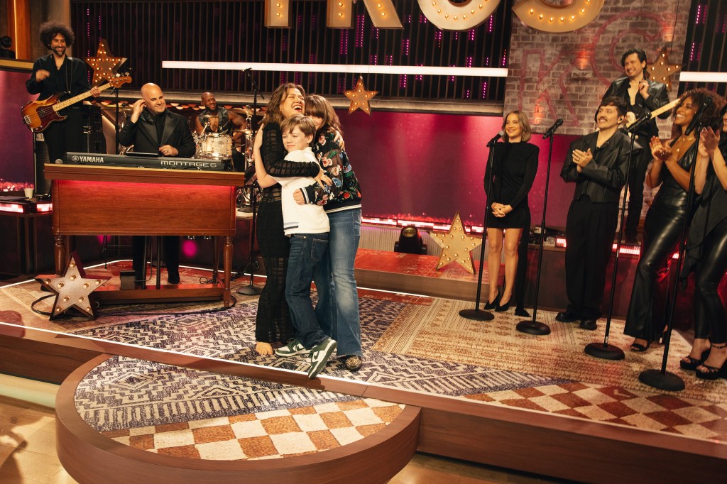 Kelly Clarkson hugs Remi Blackstock and River Blackstock on stage, surrounded by band members and guests.