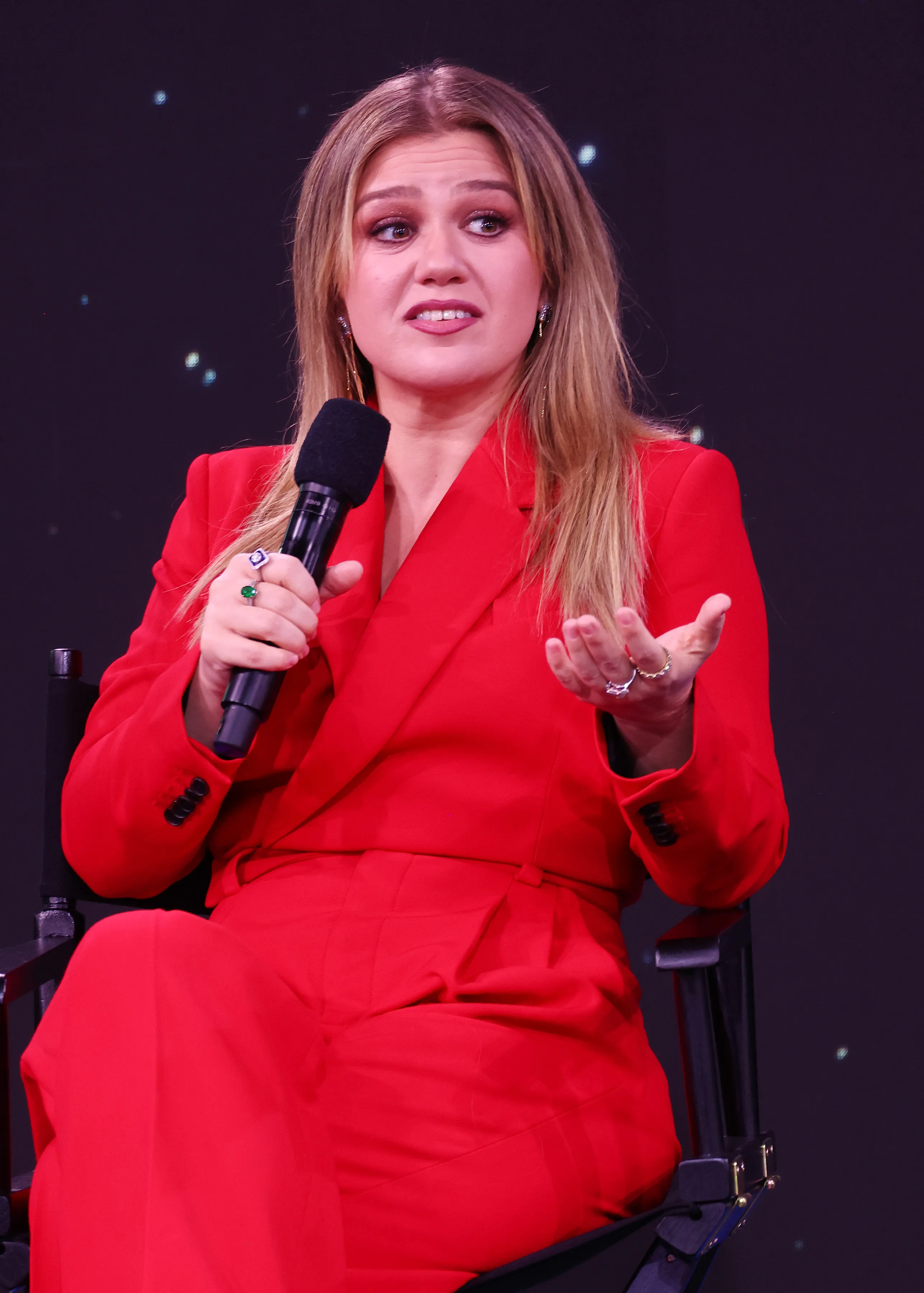 Kelly Clarkson speaks onstage in a red suit, holding a microphone.