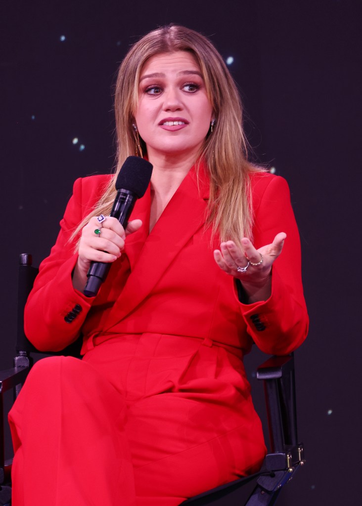 Kelly Clarkson speaks onstage wearing a red suit.